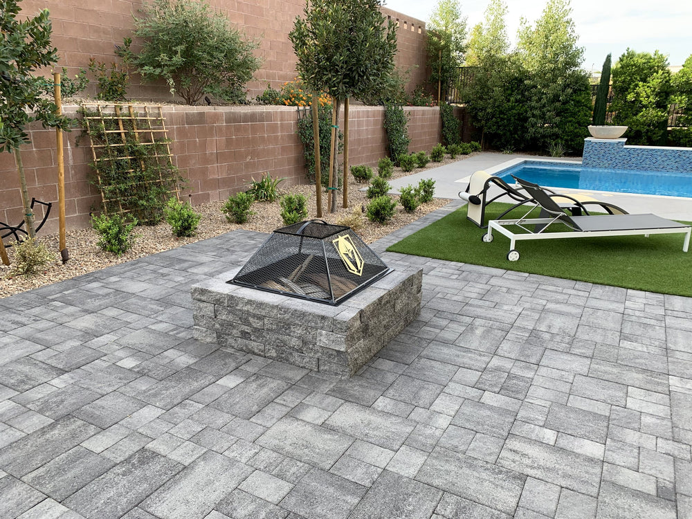 Gray paver patio with a stone fire pit, decorative rock beds and artificial turf beside a backyard pool